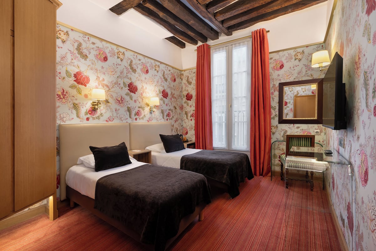 Ein Hotelzimmer mit zwei Betten, einem Holzschrank, einem Schreibtisch mit Stuhl, einem Spiegel und blumigem Wandpapier.