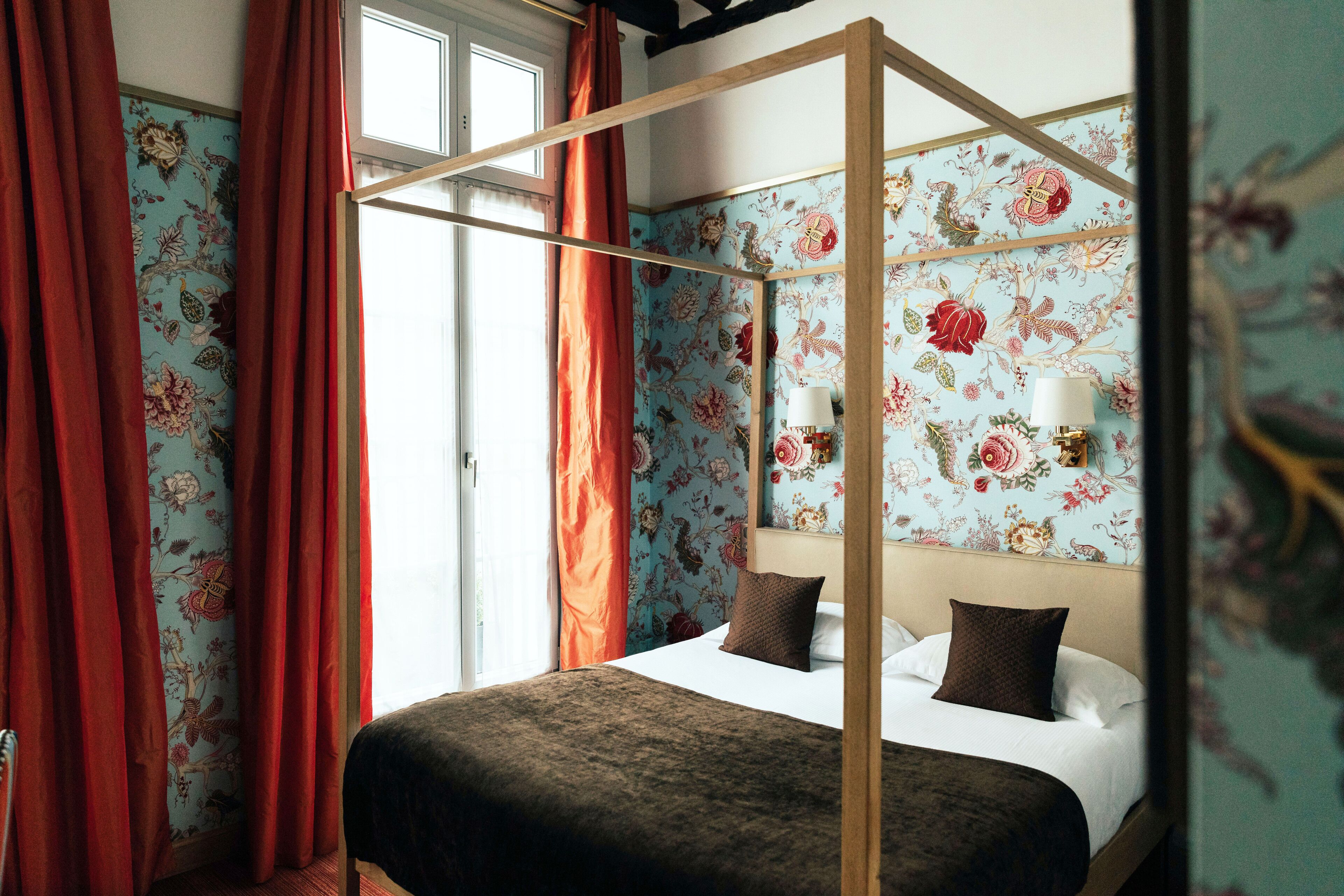A four-poster bed with floral wallpaper, red curtains, and a window with a view.
