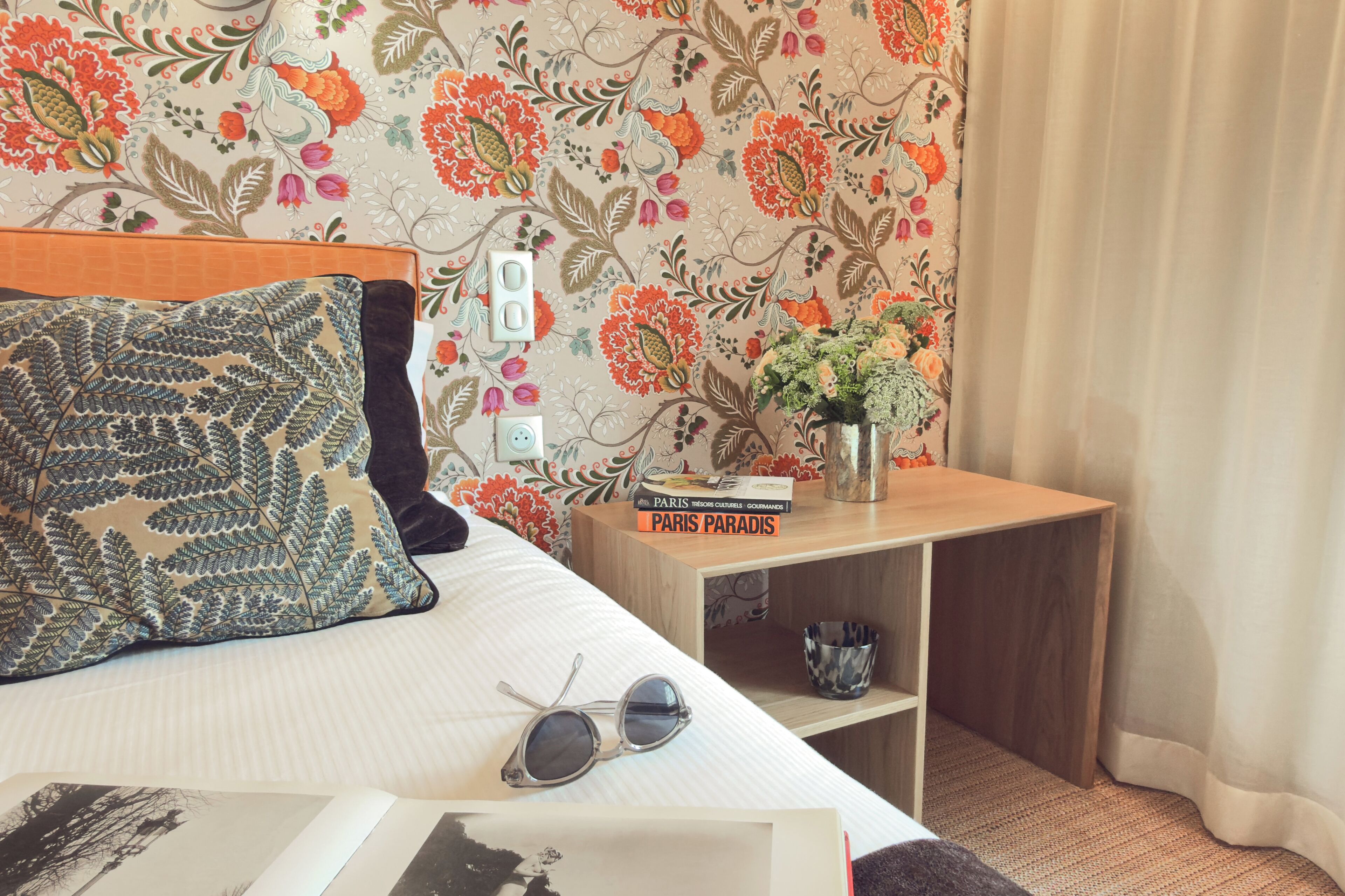 Un lit bien fait, agrémenté d’un coussin à motifs, d’une paire de lunettes de soleil et d’un livre intitulé « Paris Paradis » posé sur une table de chevet.