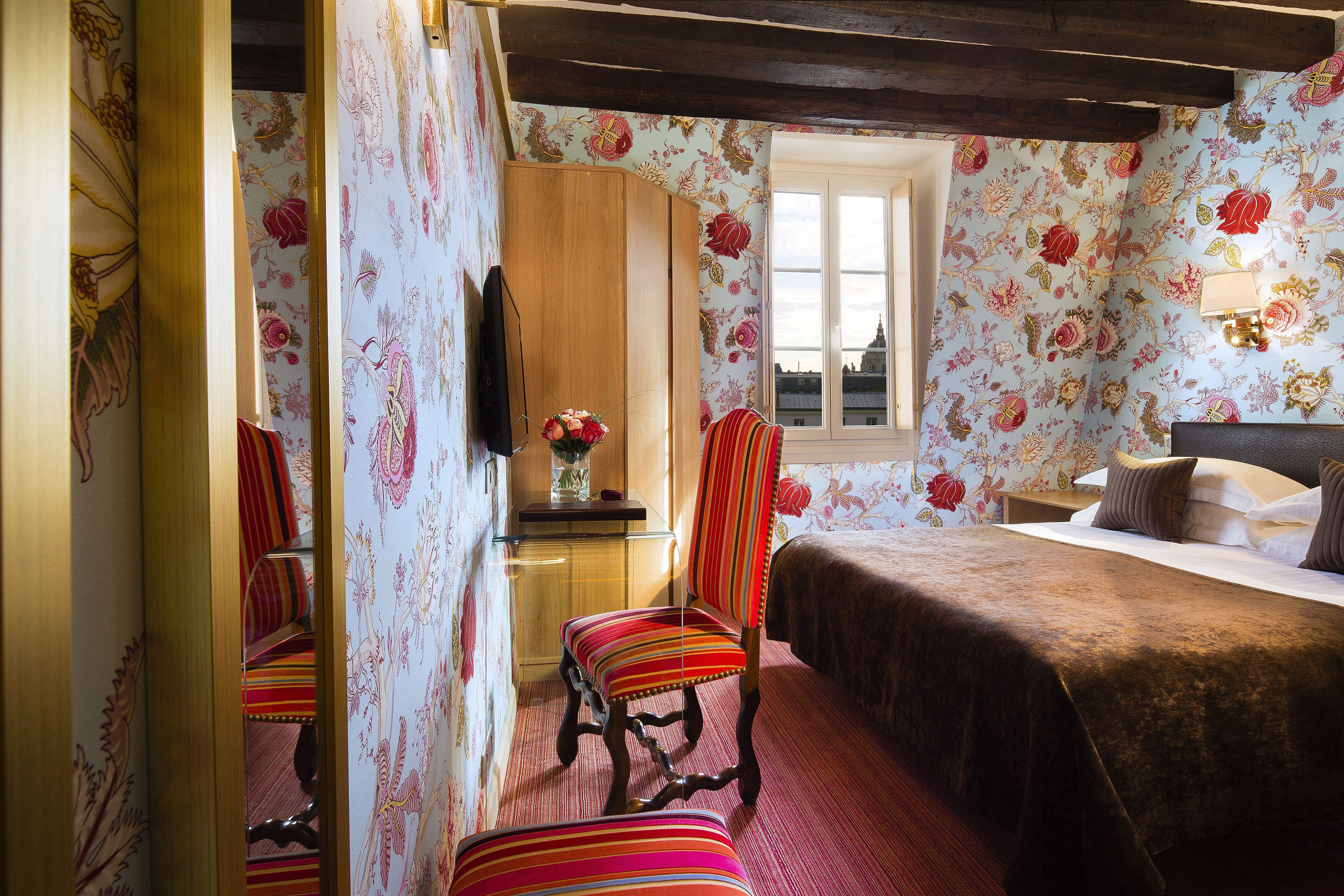 Een slaapkamer met bloemenbehang, een houten garderobe, een bed met een bruine plaid en een raam met uitzicht op gebouwen.