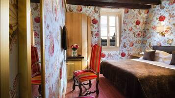 Een slaapkamer met bloemenbehang, een houten garderobe, een bed met een bruine plaid en een raam met uitzicht op gebouwen.