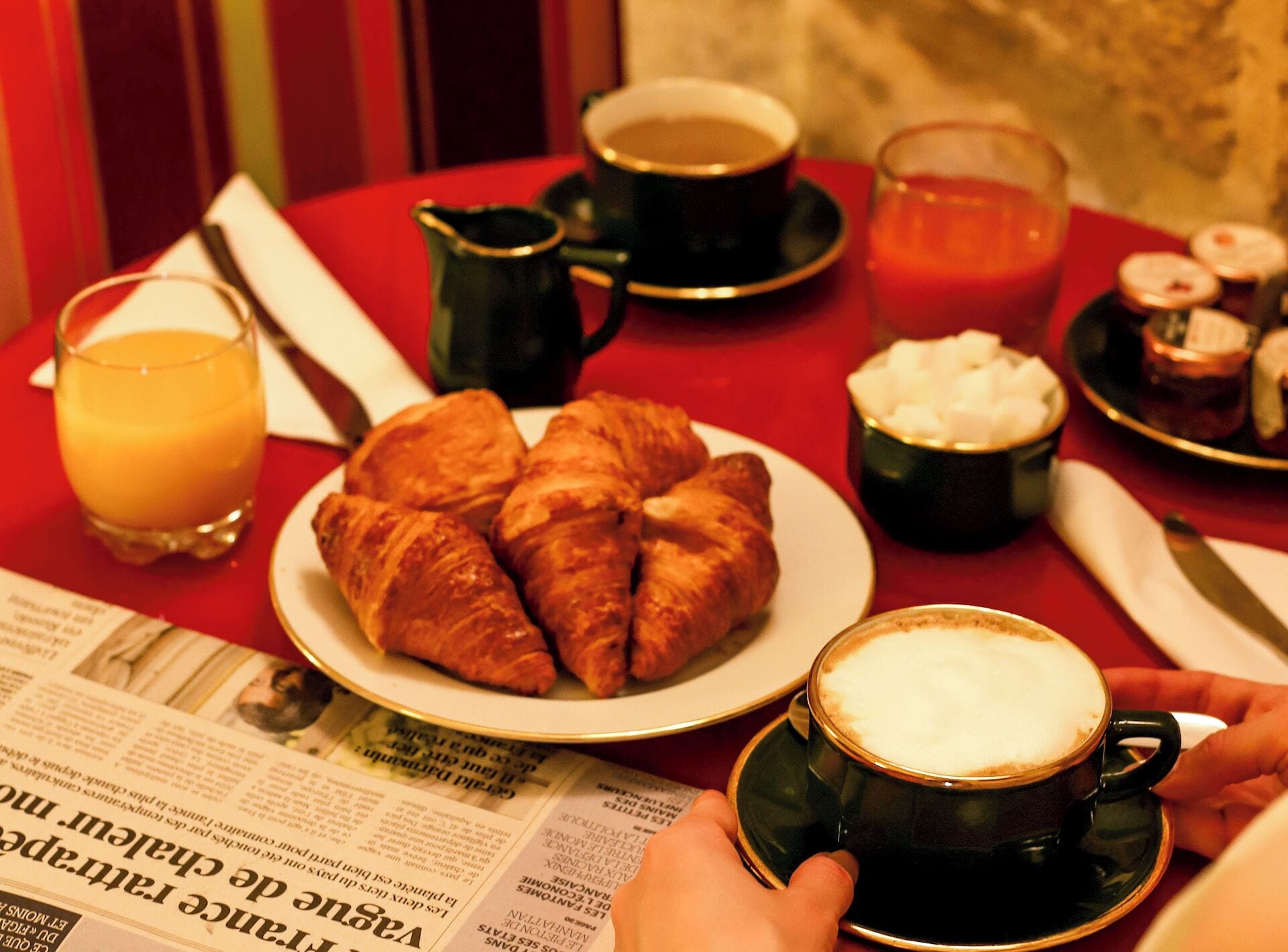 Un desayuno con cruasanes, café, jugo de naranja y un periódico sobre una mesa roja.