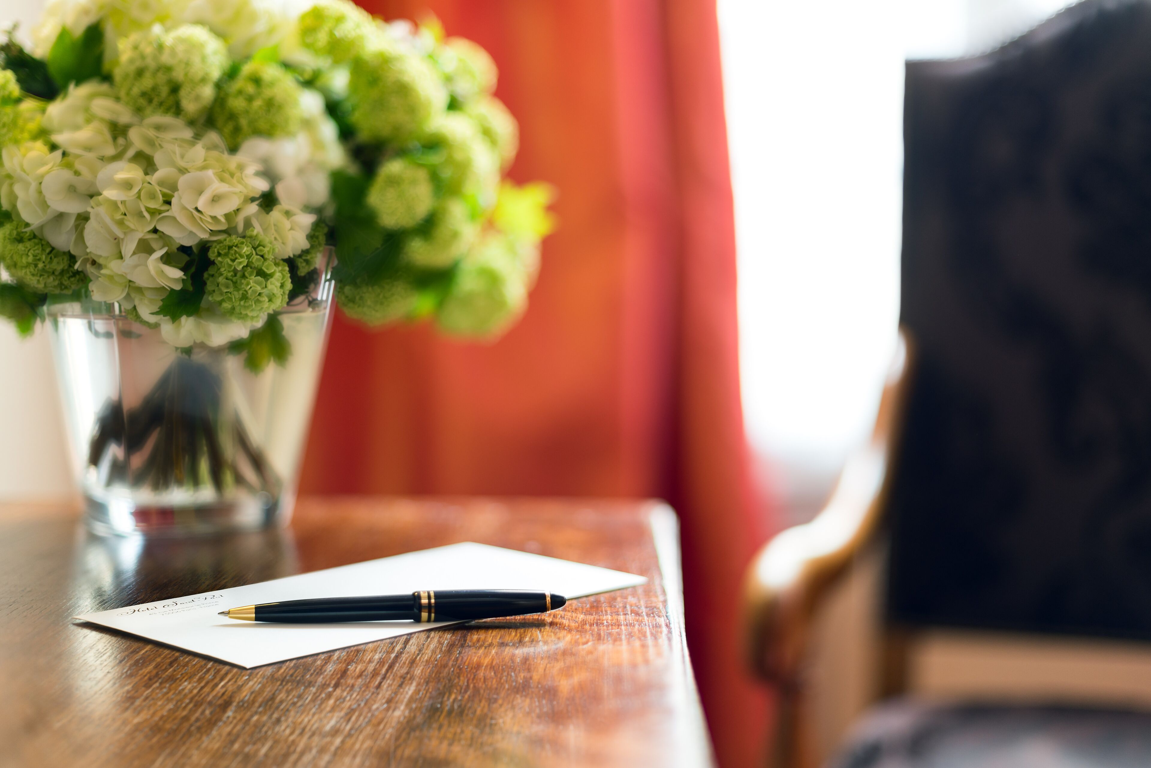 Une table en bois sur laquelle se trouvent un carnet blanc, un stylo noir et un vase avec des fleurs blanches et vertes.