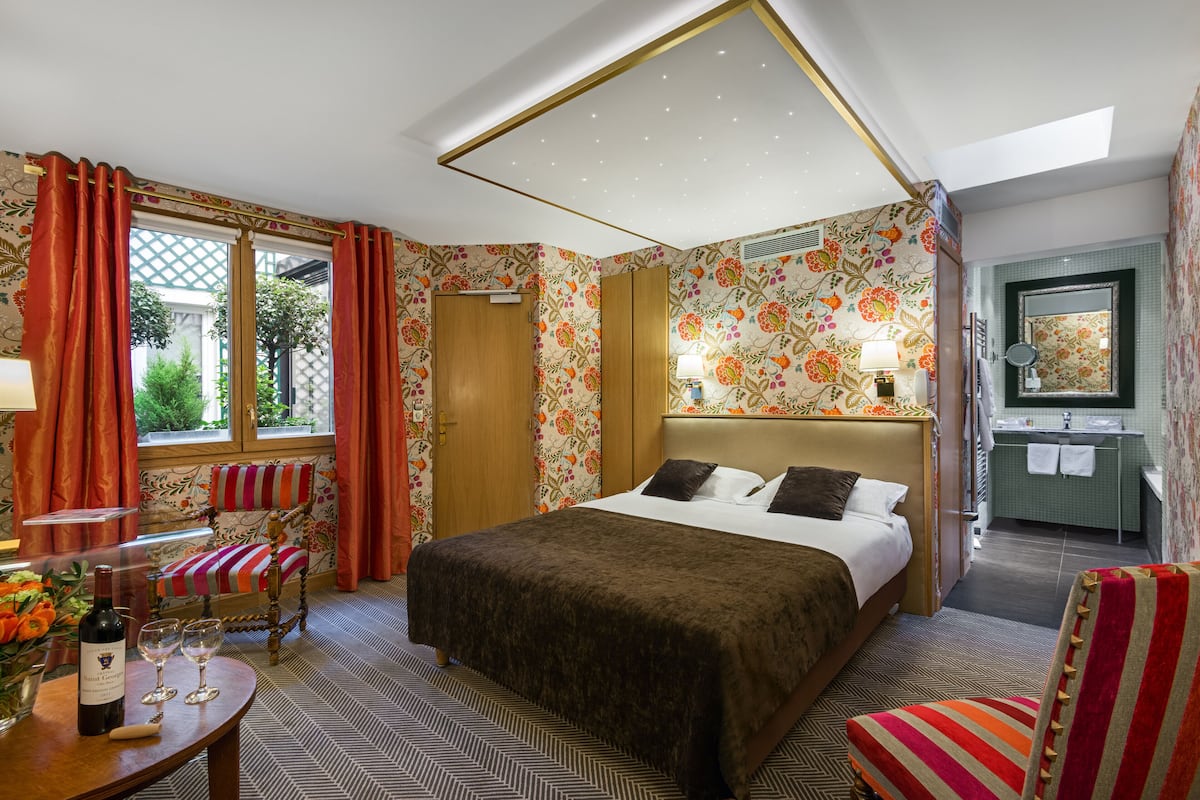 Ein Hotelzimmer mit einem großen Bett, einer Sitzecke mit einem Sessel, einem kleinen Tisch mit Flasche und Gläsern, einem Fenster mit Vorhängen und einem Badezimmer mit Spiegel und Waschbecken.