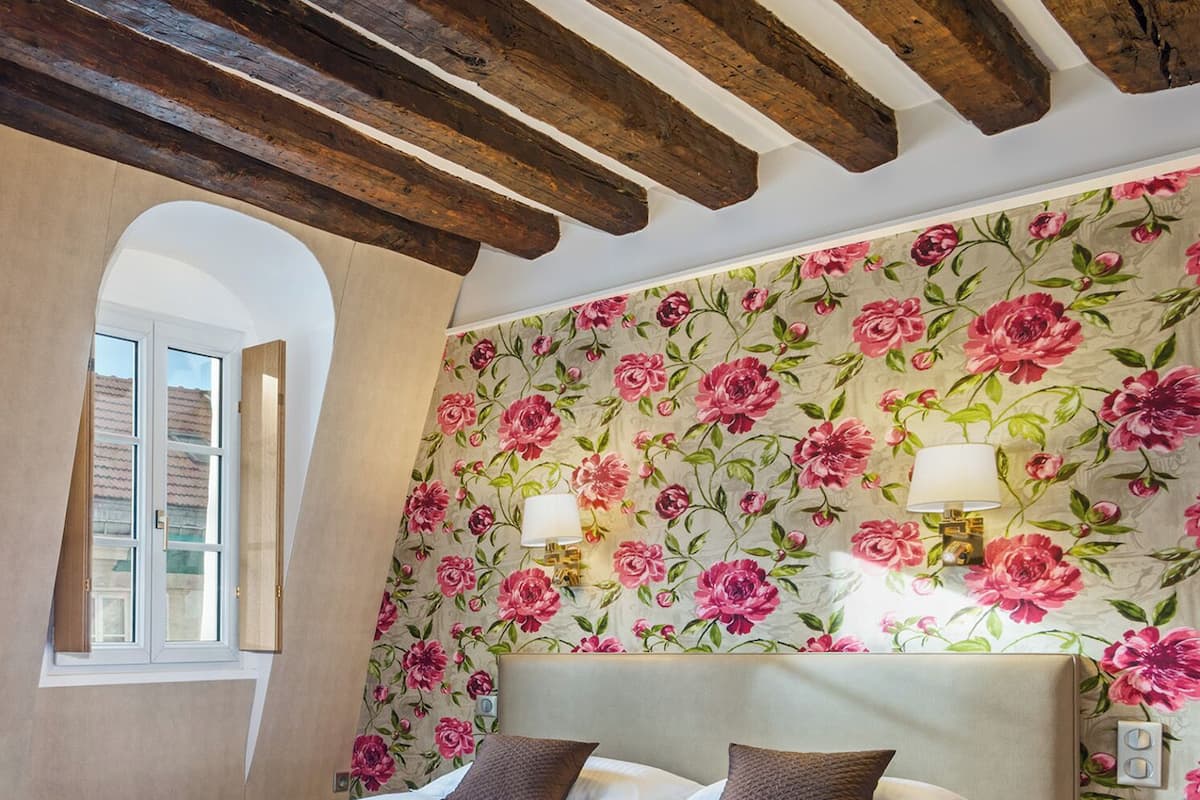 Ein Schlafzimmer mit einem Bett, blumigem Wandpapier, einem Fenster und einer Holzbalkendecke.