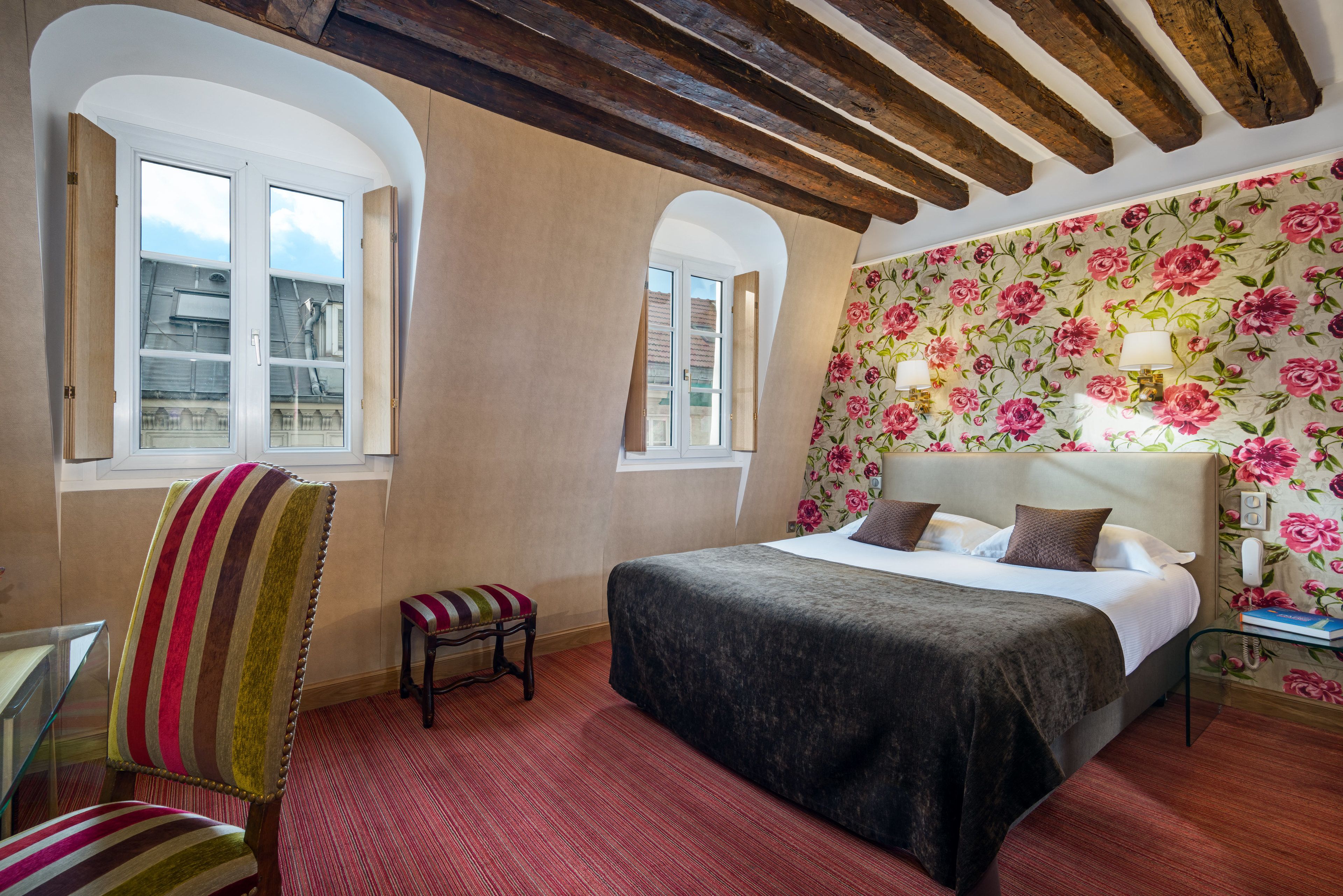 A bedroom with a large bed, a striped chair, a small stool, and a window with a view of buildings.