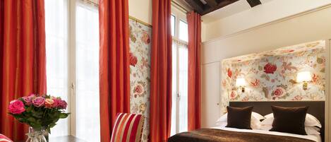 Habitación de hotel con una cama, una silla, una mesa con un florero y una ventana con cortinas.