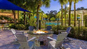 An outdoor seating area with Adirondack chairs around a fire pit, a pool in the background, and a pergola with umbrellas. - Marriott's Royal Palms (Orlando)