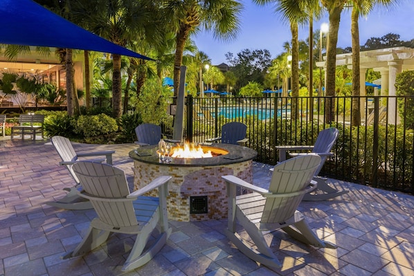 Un espace extérieur aménagé avec des fauteuils Adirondack disposés autour d’un foyer, une piscine en arrière-plan et une pergola ombragée de parasols.