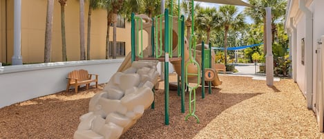 A playground with a slide, climbing structures, and a shaded area with a wooden chair.