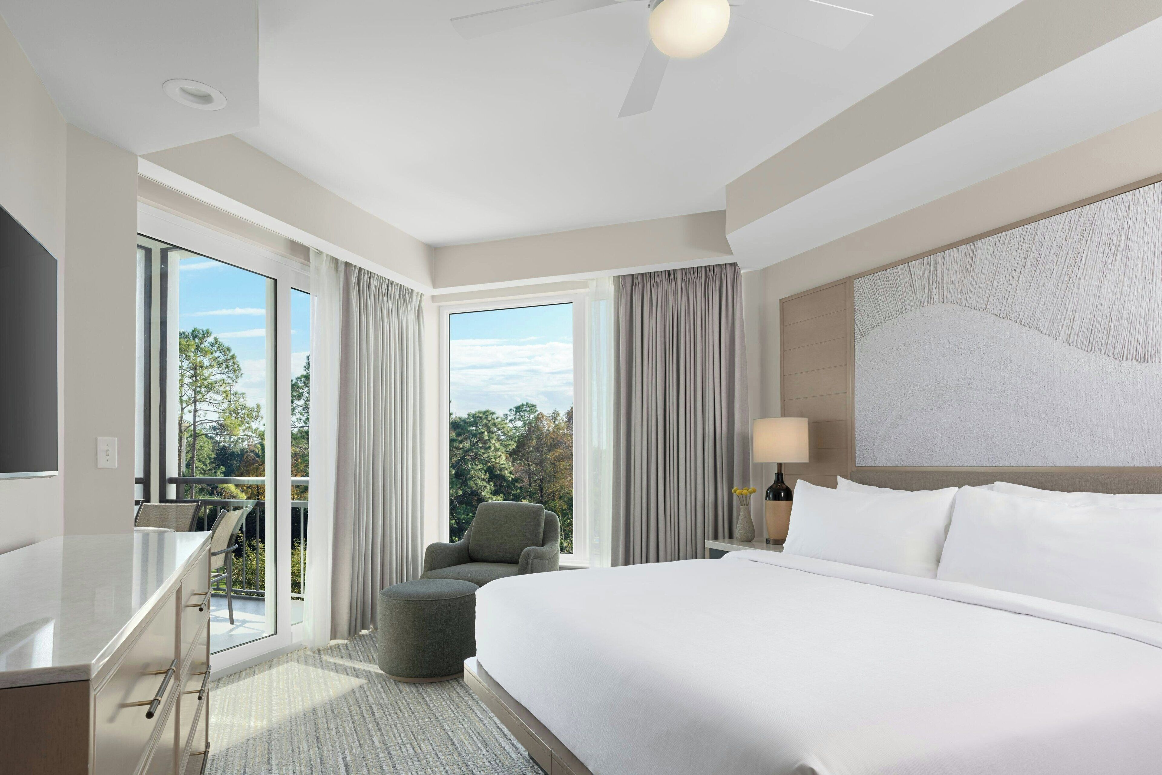 A hotel room with a large bed, a desk, a chair, and a balcony with a view of trees.