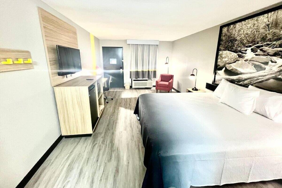 A hotel room with a bed, a desk with a chair, a TV, a window with curtains, and a painting on the wall.