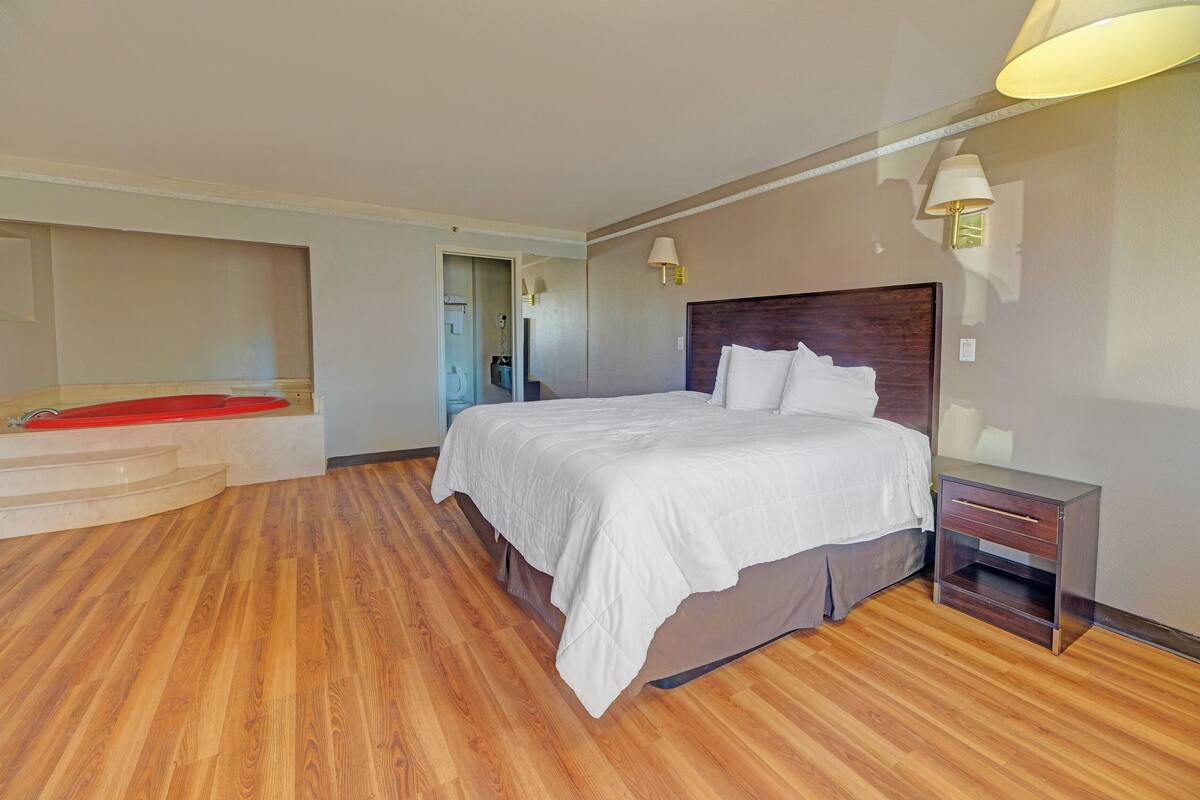 A hotel room with a large bed, wooden flooring, a nightstand, and a red chair.