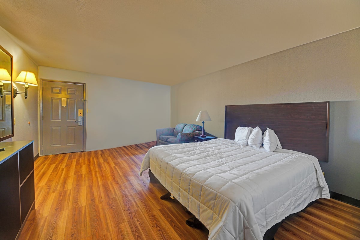 A hotel room with a wooden floor, a bed with white bedding, a chair, and a desk with a lamp.