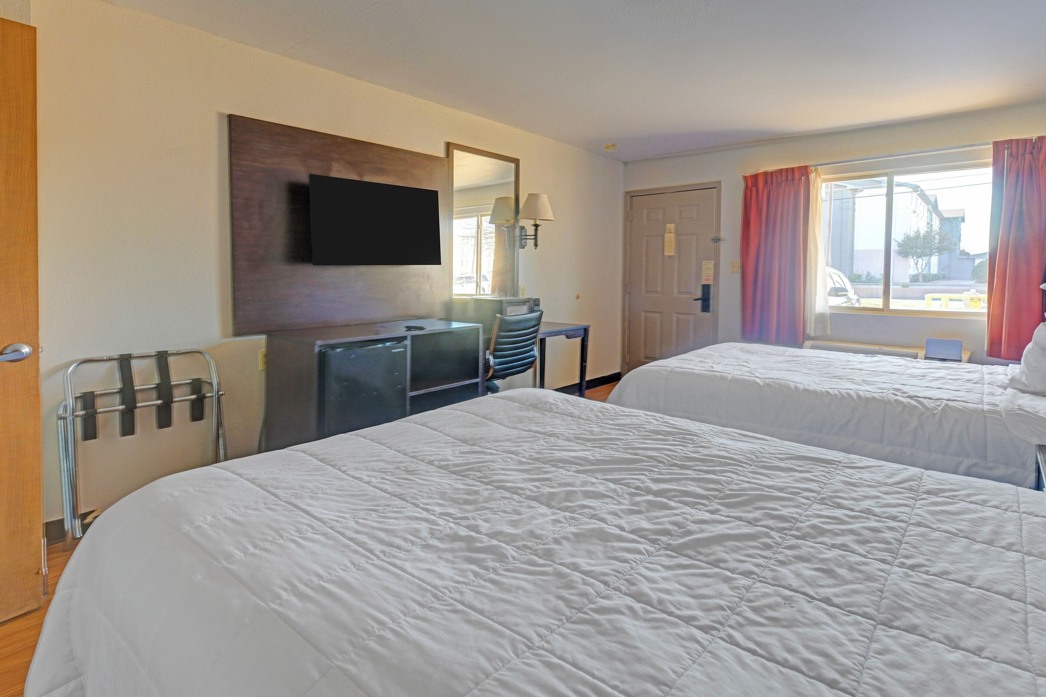 Une chambre d’hôtel avec deux lits, un bureau, une chaise, un téléviseur fixé au mur et une fenêtre avec des rideaux.