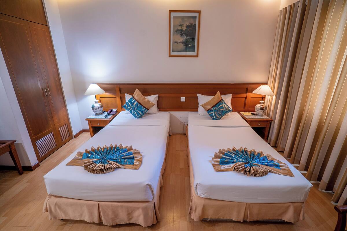 Een hotelkamer met twee bedden, een houten hoofdbord, nachtkastjes met lampen, een schilderij aan de muur, en een raam met gordijnen.