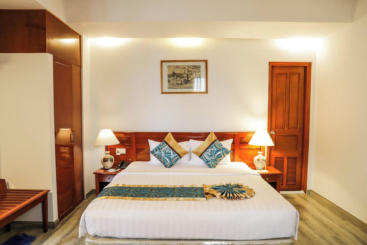 Hotelkamer met een groot bed, houten hoofdeinde, twee bedlampen, een nachtkastje met een vaas en een lijst aan de muur.