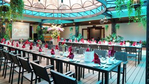 Un vaste espace intérieur aménagé pour les repas, avec de longues tables dressées pour un événement formel, entouré d’une végétation luxuriante et éclairé par des luminaires modernes.