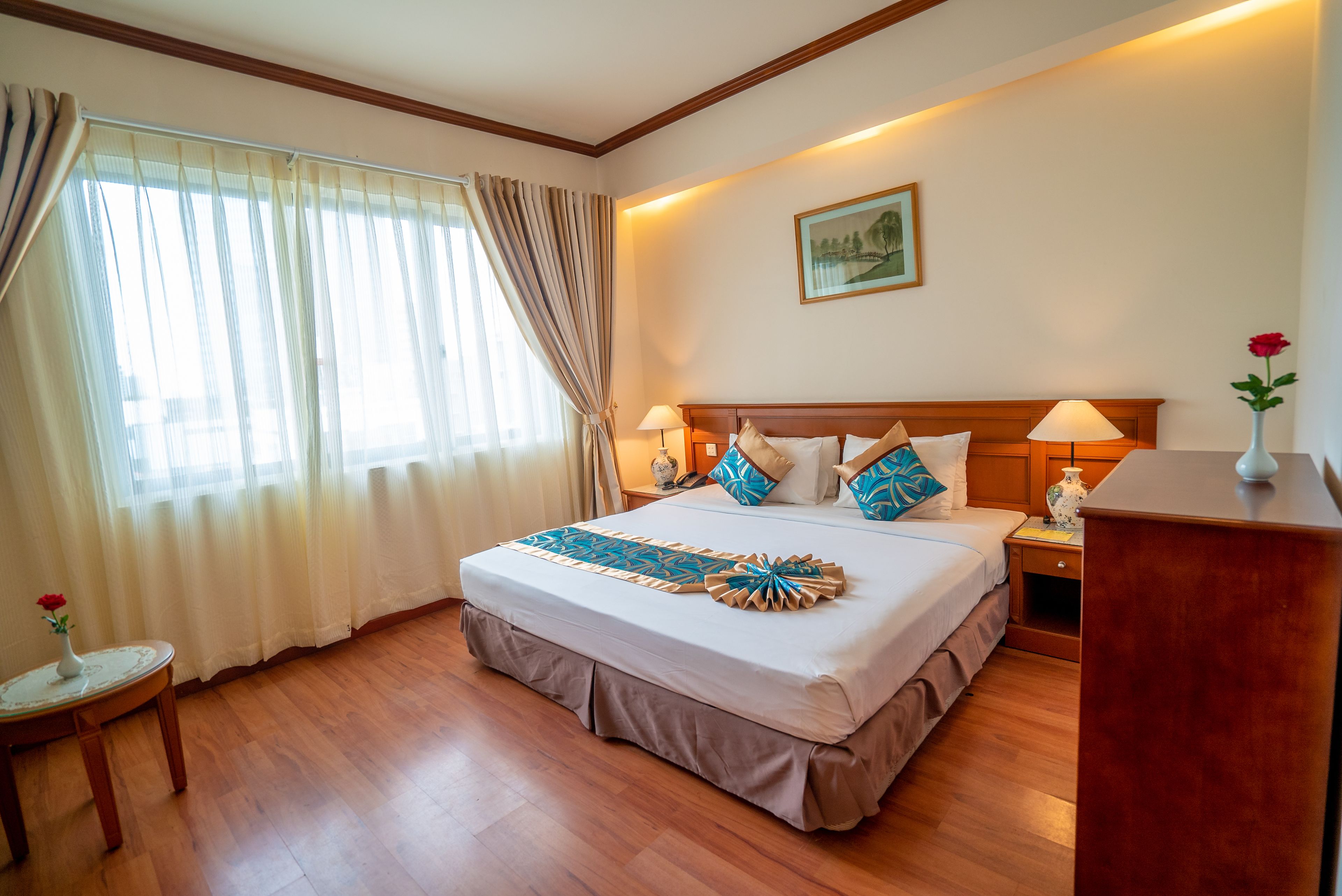 A hotel room with a large bed, a bedside table, a nightstand, a desk, a chair, a window with curtains, and a painting on the wall.