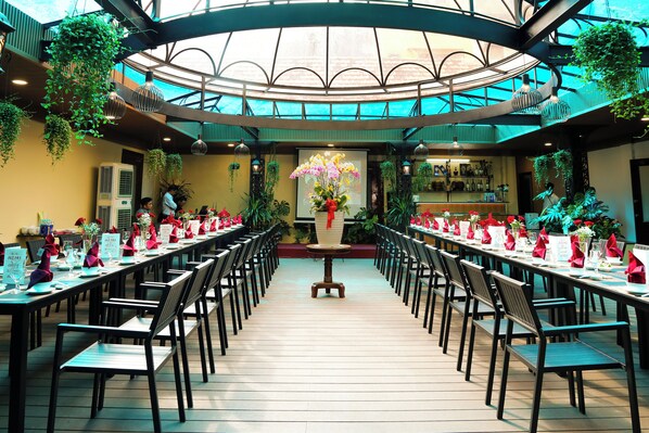 A long dining table with chairs and a central floral arrangement in a spacious, well-lit room with a high ceiling and hanging plants.