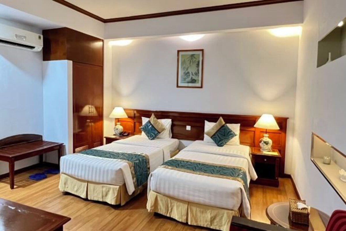 Une chambre d’hôtel avec deux lits, des têtes de lit en bois, des lampes de chevet, une petite table et un climatiseur fixé au mur.