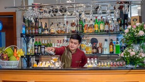 Un espace bar avec un barman préparant un cocktail, des étagères remplies de différentes bouteilles d’alcool et une composition florale décorative.