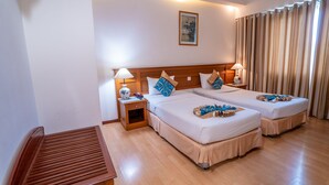 Une chambre d’hôtel avec deux lits, des têtes de lit en bois, des tables de chevet avec des lampes, un petit banc en bois et une grande fenêtre avec des rideaux.