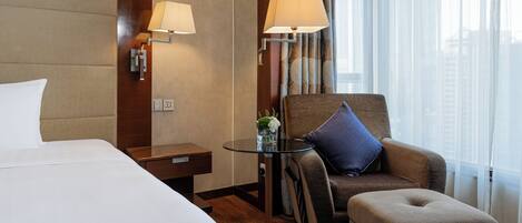 A hotel room with a large bed, a small round table with a lamp, a sofa, and a chair.