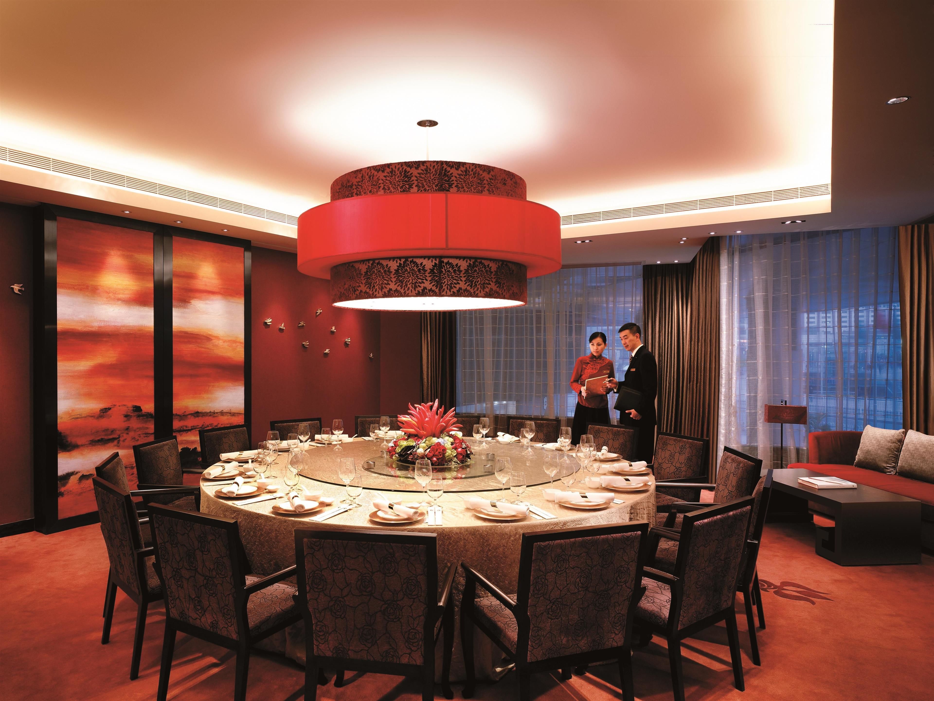 Une salle à manger d’hôtel avec une table ronde dressée pour un repas, entourée de chaises, et un grand lustre rouge.