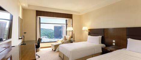 A hotel room with two beds, a desk, a chair, a lamp, and a view of the city.