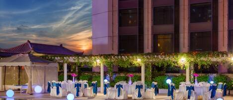 An outdoor wedding setup with tables and chairs, a pool, and a building in the background.