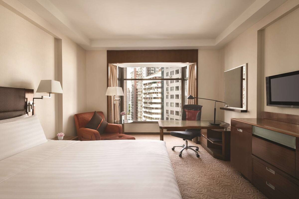 A hotel room with a large bed, a desk, a chair, a television, and a window with a city view.