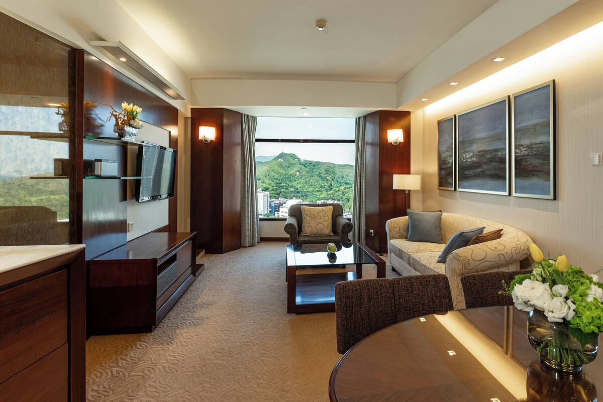 A modern hotel room with a large window offering a mountain view, a flat-screen TV, a sofa, a coffee table, and a dining table.