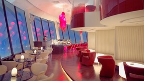 A modern bar area with a curved counter, seating with red and white chairs, and large windows with city views. - Shangri-La Shenzhen (Shenzhen)