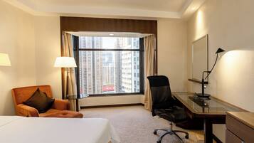 Habitación de hotel con cama, escritorio, silla, lámpara y una ventana grande con vista a la ciudad.