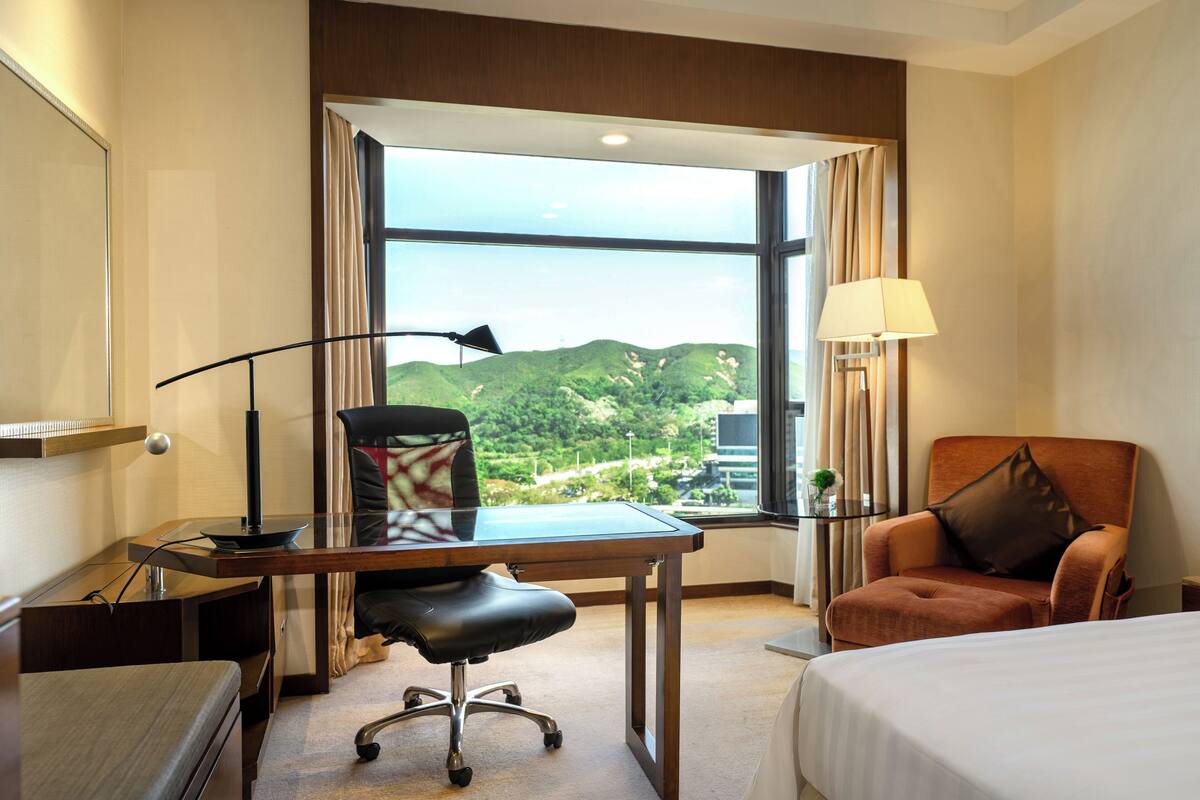 A hotel room with a desk, chair, bed, and a large window offering a view of greenery and buildings.