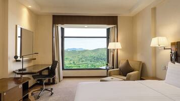 A hotel room with a large bed, a desk with a chair, a small table, and a view of green hills through a large window.
