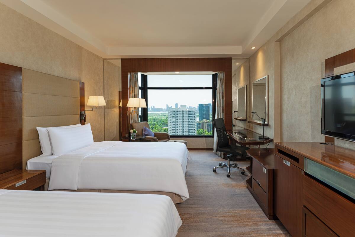 A hotel room with two beds, a desk, a chair, a television, and a large window offering a city view.