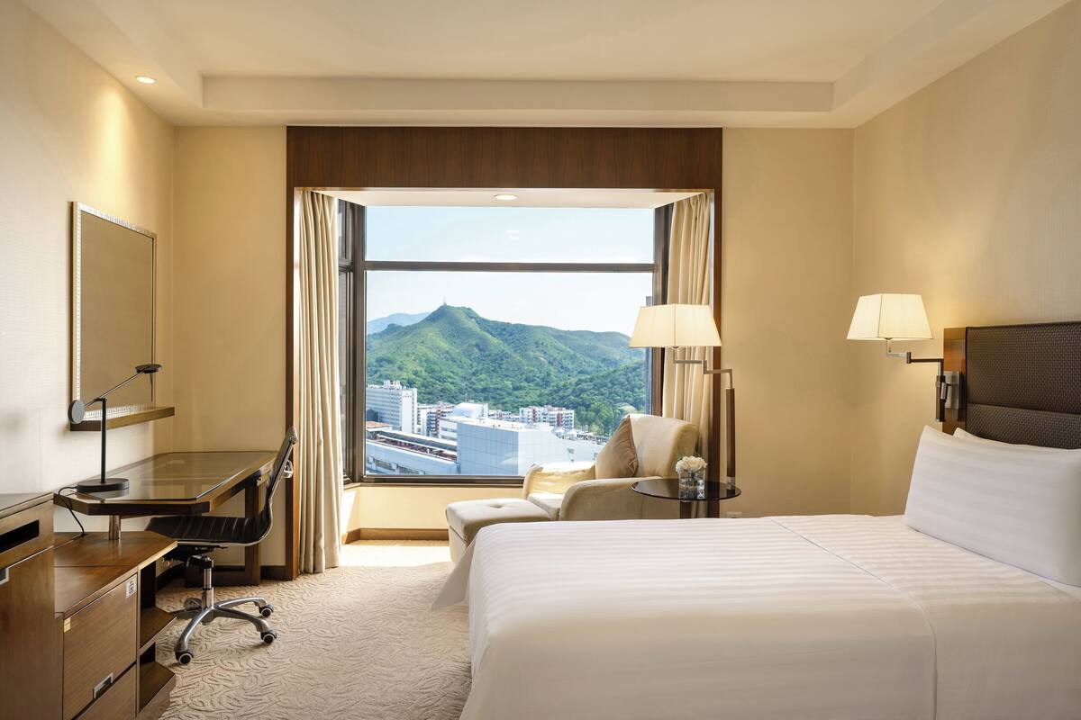 A hotel room with a large bed, a desk with a chair, a small table with a lamp, and a view of a city and mountains.