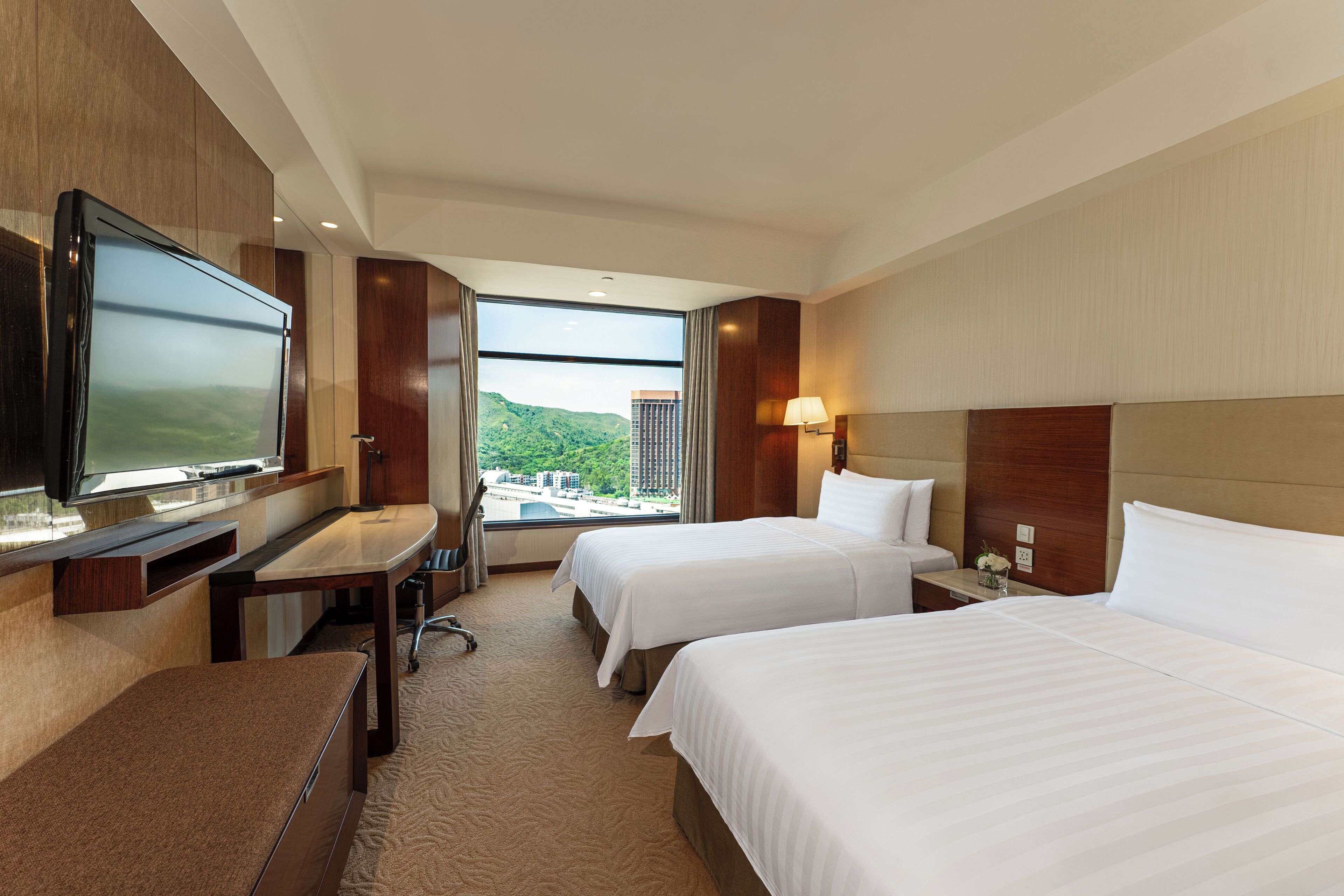 A hotel room with two beds, a desk, a TV, and a window with a view of a cityscape and mountains.