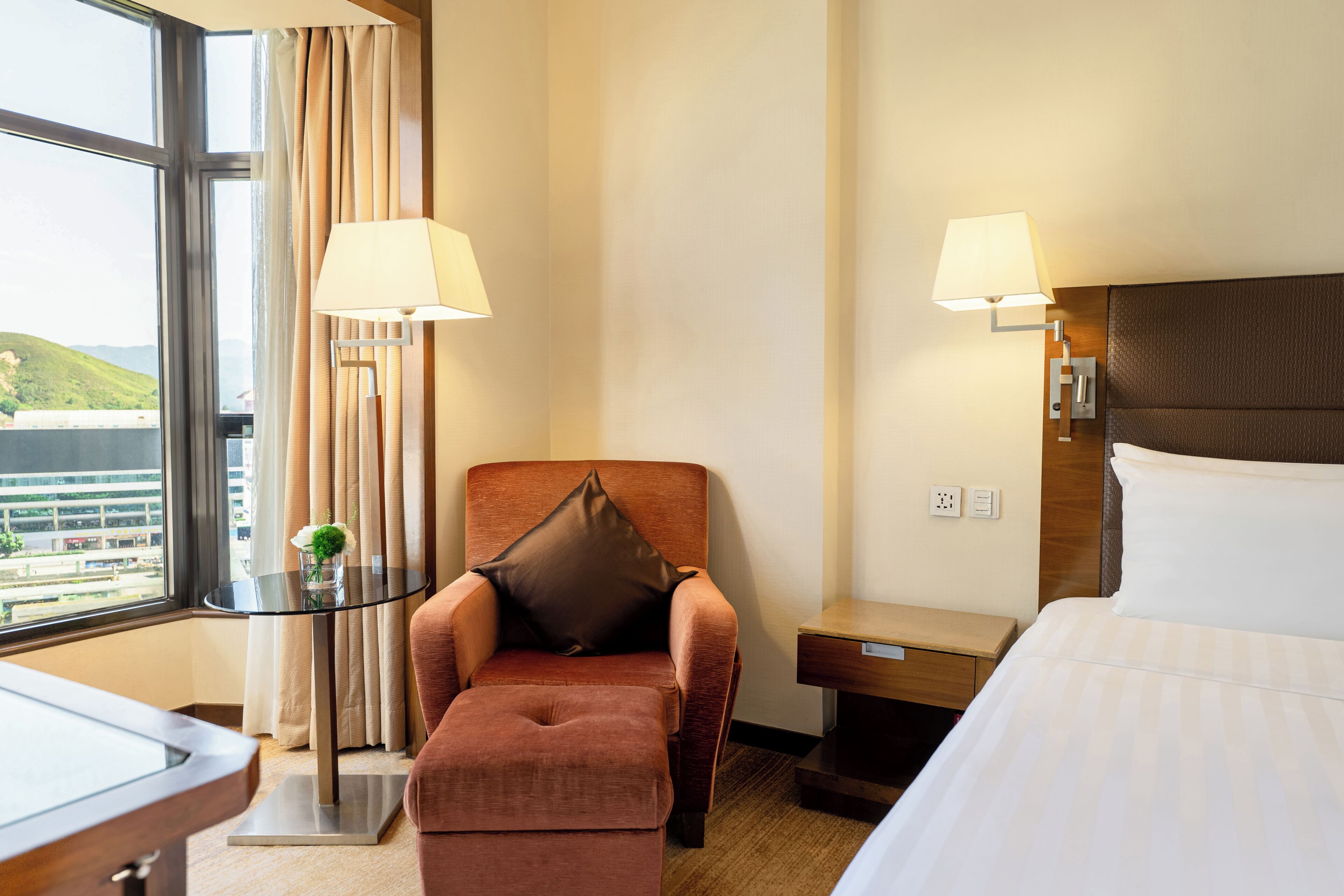A hotel room with a bed, a chair with a pillow, a small table with a lamp, and a view of the city through a window.