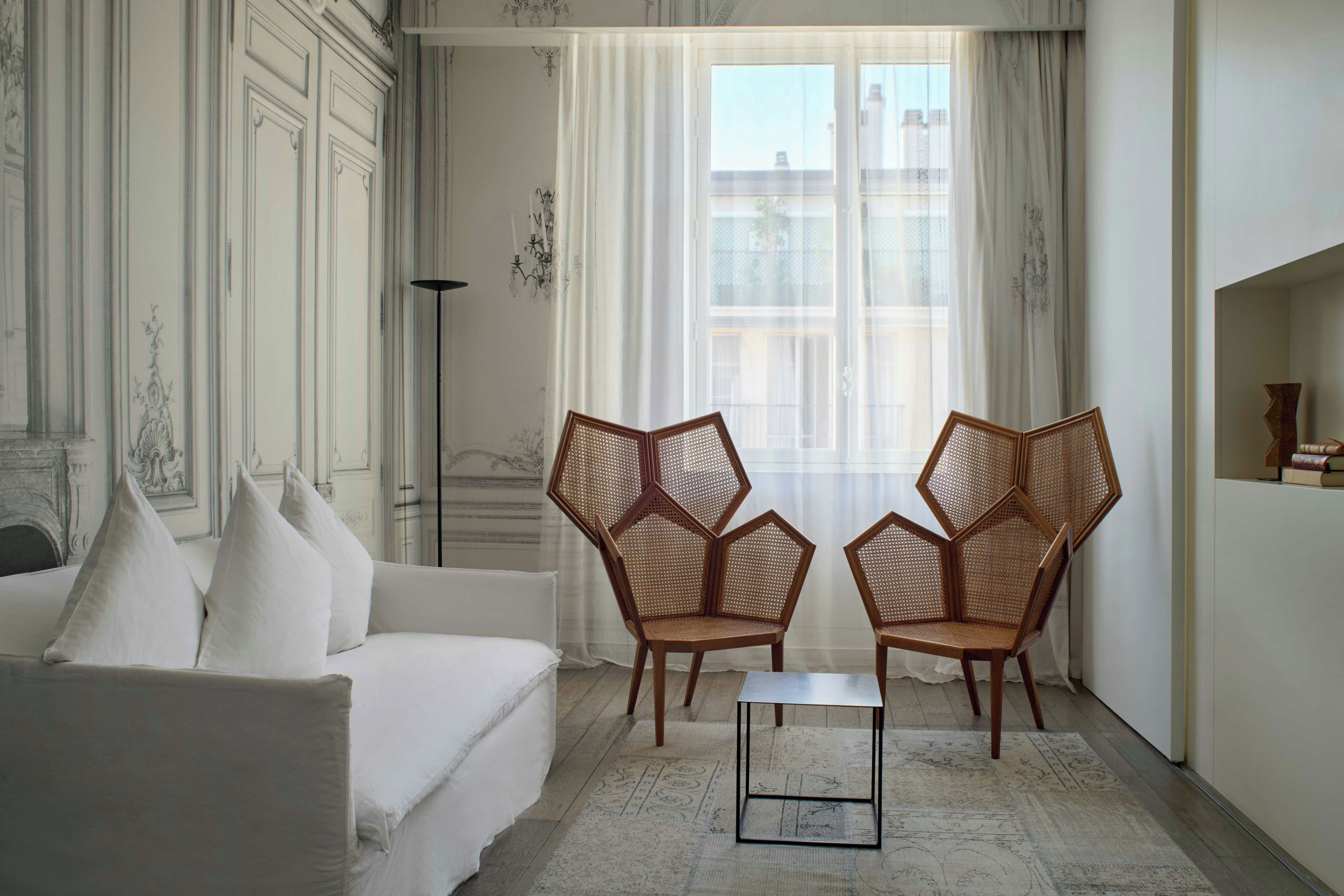 A room with a white sofa, two hexagonal wicker chairs, a small table, and a large window with sheer curtains.