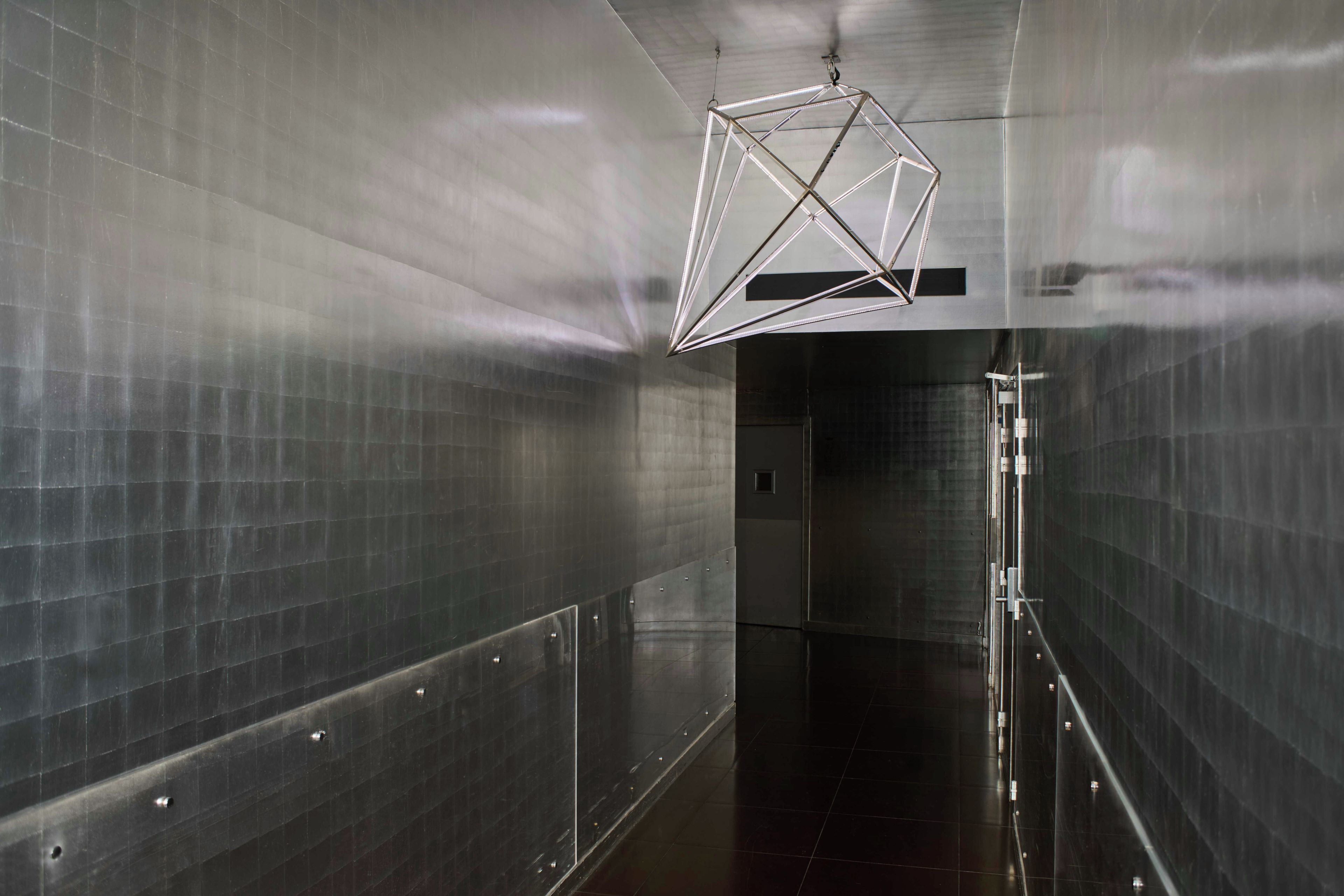 Un couloir aux murs métalliques réfléchissants et doté d’un luminaire aux formes géométriques.