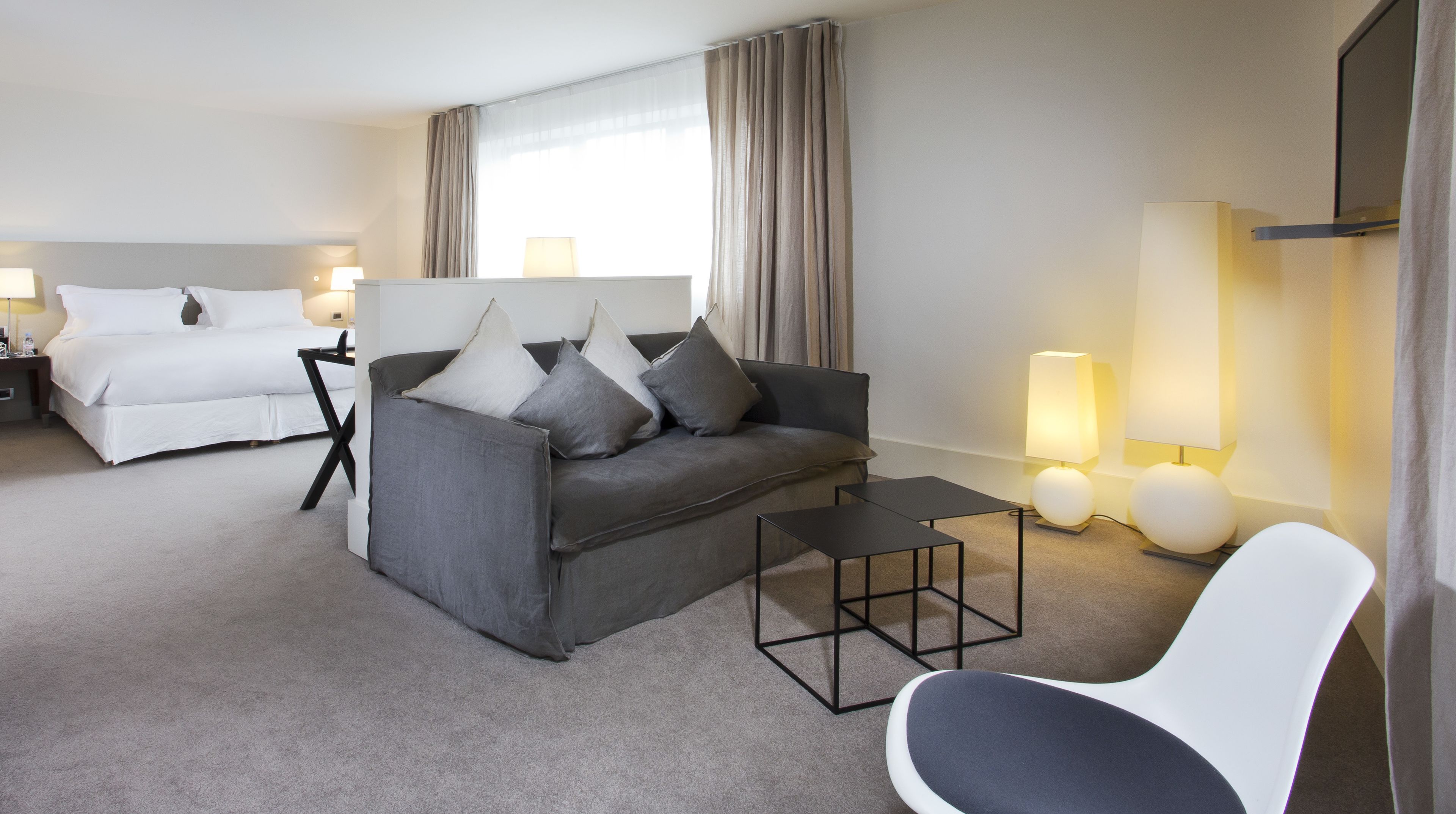 A hotel room with a bed, a grey sofa with pillows, a small table, a white chair, and two bedside lamps.