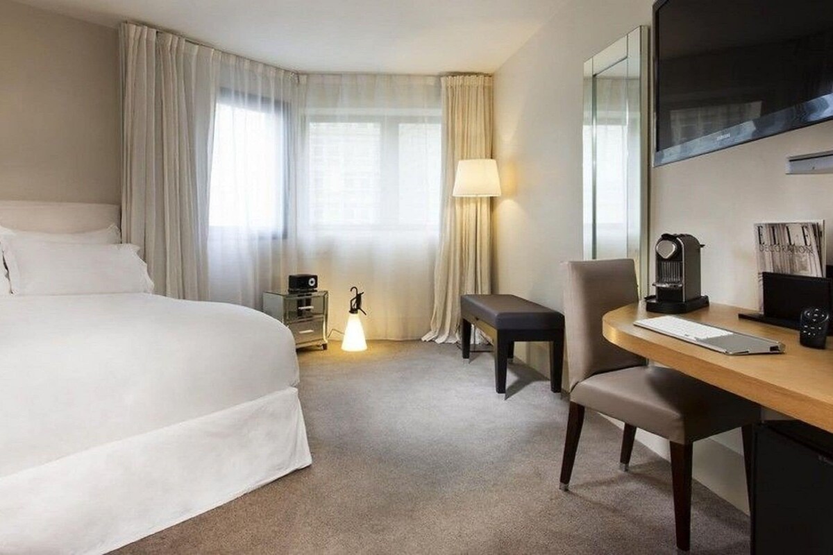A hotel room with a large bed, a desk with a chair, a lamp, a coffee machine, and a TV.