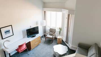 A modern hotel room with a flat-screen TV, a desk with a red chair, a grey sofa, and a large window with curtains.