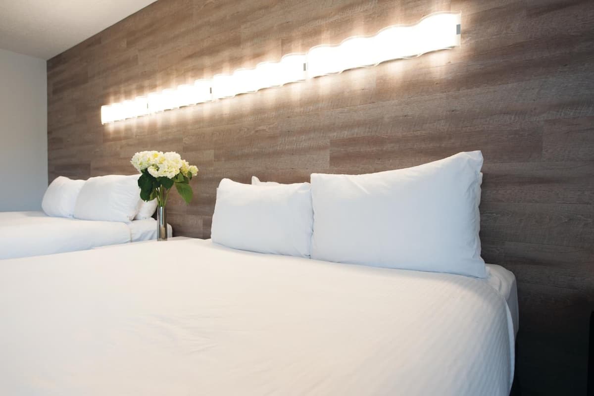 Ein Hotelzimmer mit einem großen Bett, zwei weißen Kissen, einer Vase mit Blumen und einer Holzwand mit Lichtleiste.