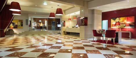 A hotel lobby with a checkered floor, red pendant lighting, and a reception desk.