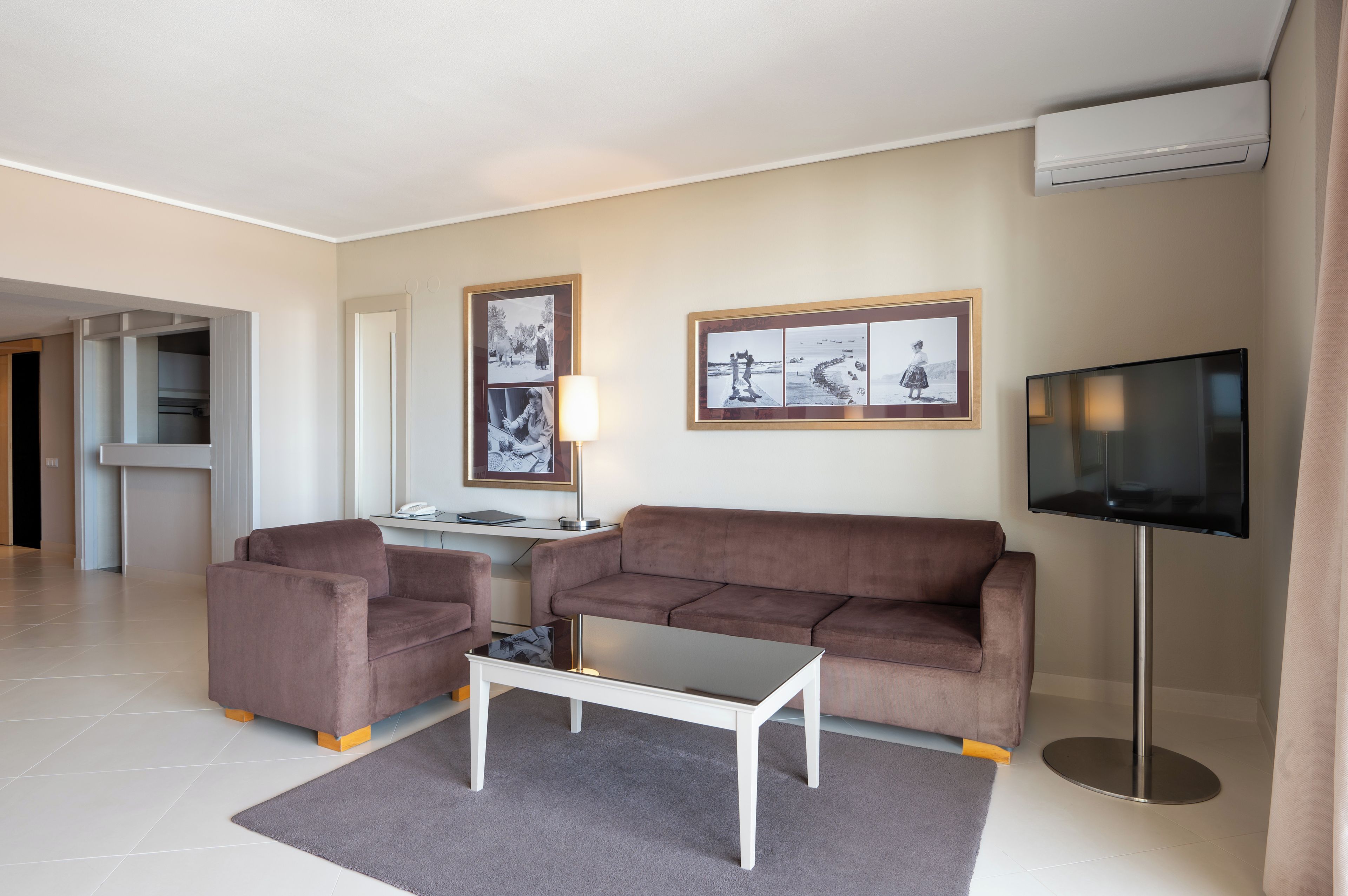 Un salon moderne avec un canapé, des fauteuils, une table basse et un téléviseur à écran plat. Des tableaux encadrés ornent le mur, et il y a un bureau avec une lampe.