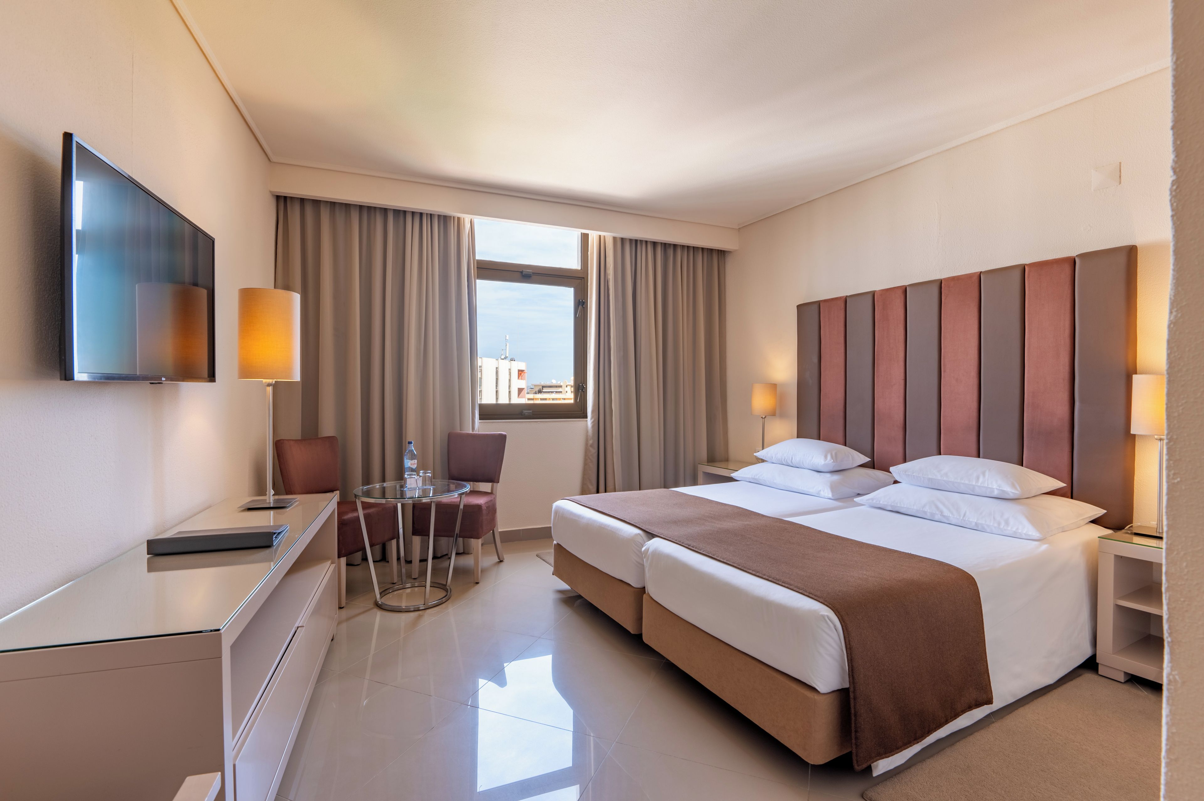 Habitación de hotel con cama, mesitas de noche, escritorio, silla, televisor y ventana con cortinas.